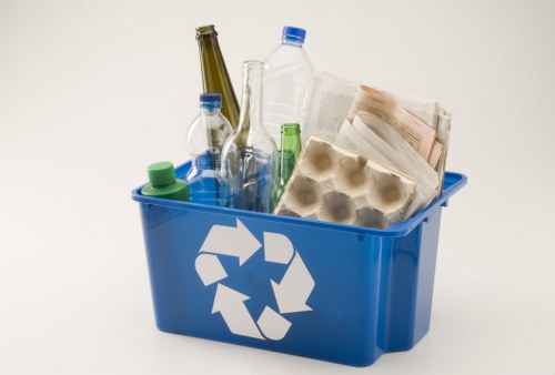 A blue recycling bin with a white recycling symbol on the side, filled with a variety of waste items including several clear plastic bottles with blue, green, and natural-colored caps, an egg carton made of cardboard with egg compartments, a crumpled newspaper or paper documents, and a tall, empty glass bottle with a dark green tint. The bin is placed on a plain white background, and the waste materials are visibly overflowing from the top of the bin, indicating a collection of general rubbish suitable for a professional waste management service in Charlton. The plastic bottles' surfaces are smooth and shiny, with some showing slight creases or folds, and the newspaper appears crumpled and folded, while the egg carton has a foam-like texture with a slightly rough surface. The overall scene emphasizes waste collection involving both recyclable plastics and paper products, relevant to local rubbish removal services offered by Commercial Waste Charlton.