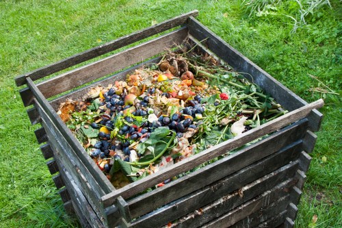 A black wooden container or compost bin positioned on a grassy area outdoors, filled with an assortment of organic waste including fruit and vegetable peelings, vegetable tops, and garden clippings. The waste displays a variety of colours and textures, from the orange and yellow of peels to the green of leaves and stems, with some soft, decomposing matter visible among the fresh garden prunings. The surrounding environment features lush, well-maintained grass, indicating a garden or outdoor space. The container shows signs of weathering, with the wood appearing slightly aged but still sturdy. This image relates to waste management services, such as those provided by Commercial Waste Charlton, focusing on organic or garden waste collection or composting, with the scene capturing the typical contents of green waste or biodegradable rubbish stored in a suitable outdoor container.
