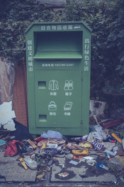 A green outdoor clothing donation and textile collection bin positioned against a backdrop of dense foliage and trees. The bin features Chinese characters indicating it is for used clothing and textil