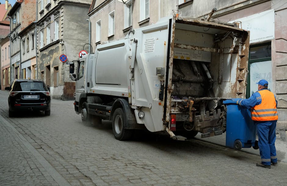 An outdoor scene showing a large amount of mixed waste and rubbish scattered on a dirt ground, with three individuals collecting debris around a white delivery truck marked with the code 'AZ 99-FX-185