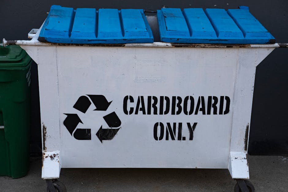 A large white wheeled bin designated for cardboard disposal, positioned outdoors on a paved surface against a dark background. The bin features a prominent black recycling symbol and the words 'CARDBO